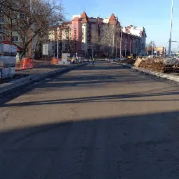 Nowa nawierzchnia asfaltowa na miejskiej ulicy w trakcie budowy, z widocznymi robotnikami w oddali i budynkami mieszkalnymi w tle.