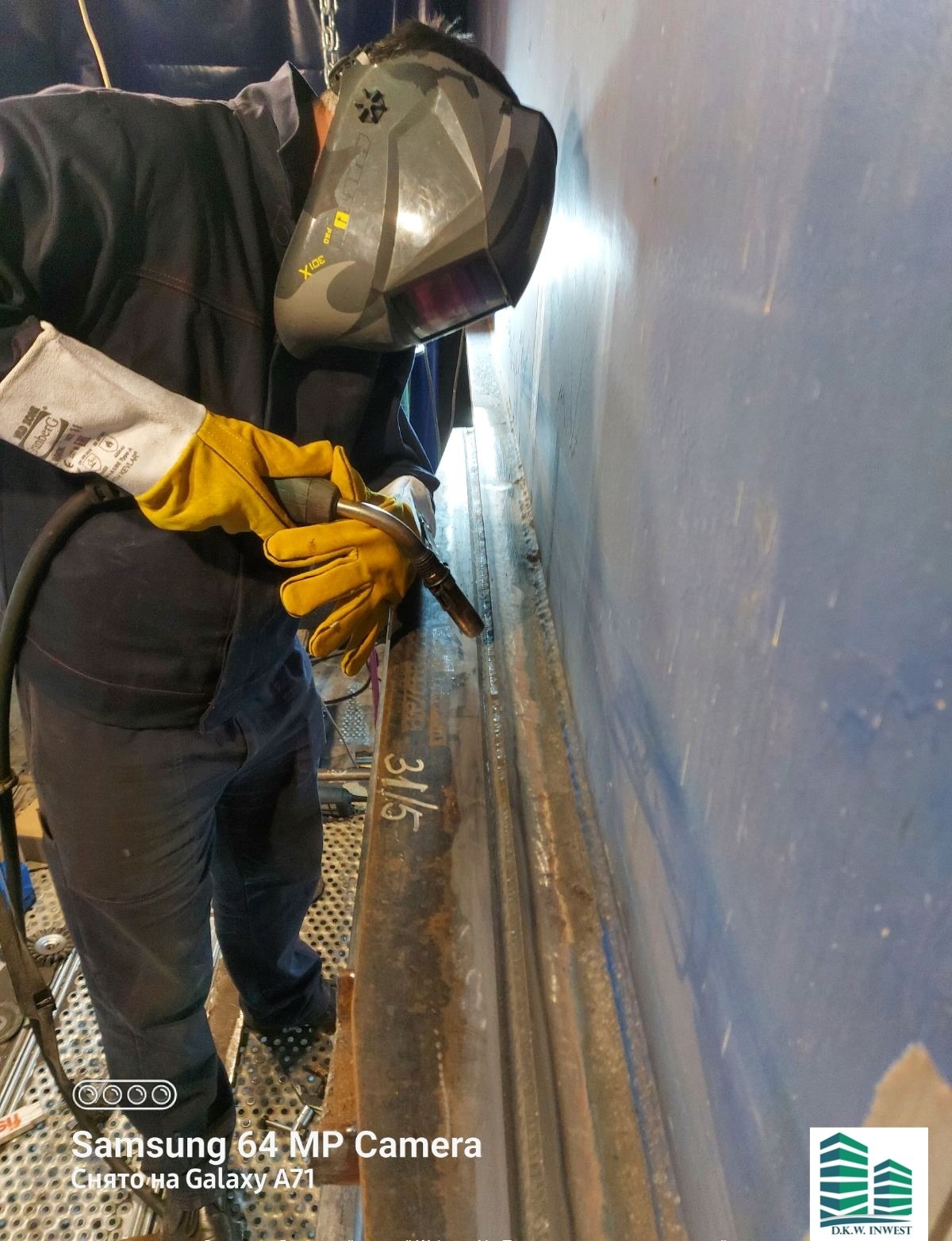 Spawacz w masce ochronnej łączy metalowe elementy konstrukcyjne, widoczne żółte rękawice ochronne i spawarka, na elemencie konstrukcyjnym widoczny napis 31/5.