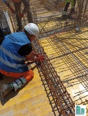 Pracownik w kasku i odblaskowej kamizelce łączy zbrojenie na drewnianym szalunku, widoczne elementy konstrukcji z prętów stalowych.