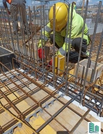 Pracownik w kasku i kamizelce odblaskowej, w trakcie montażu zbrojenia fundamentów na drewnianym szalunku.