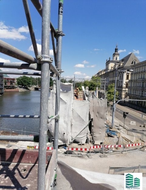 Budowa lub renowacja budynku w mieście, widok z rusztowania na plac budowy, rzekę i zabudowę miejską w tle.