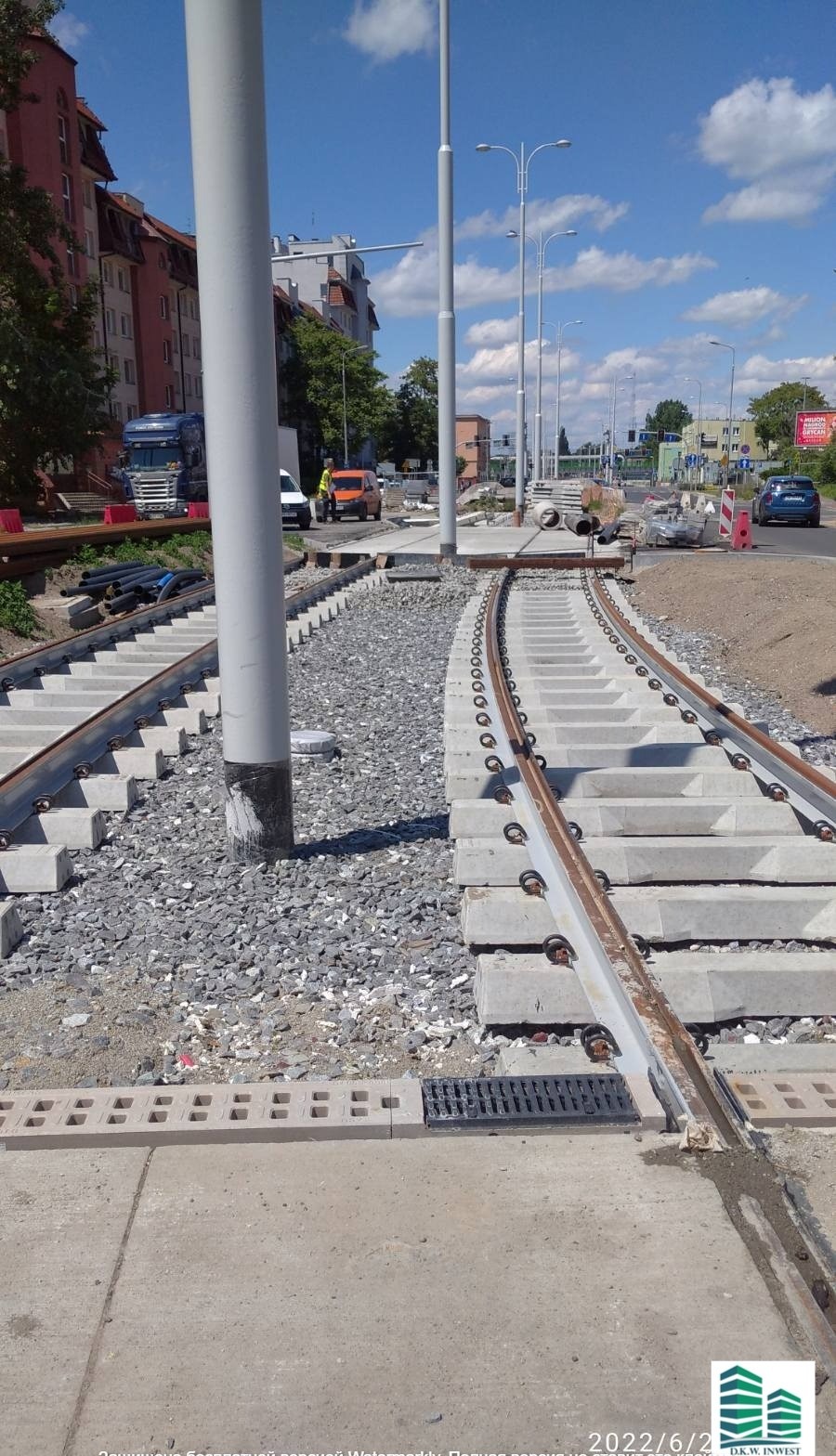 Budowa torowiska tramwajowego z betonowymi podkładami i podsypką żwirową, widoczny fragment ulicy z budynkami i pojazdami w tle.