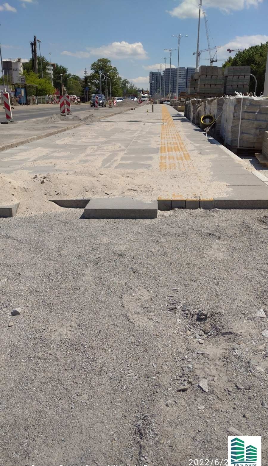 Fragment chodnika w trakcie budowy z elementami dla niewidomych, w tle widoczne budynki i dźwig.