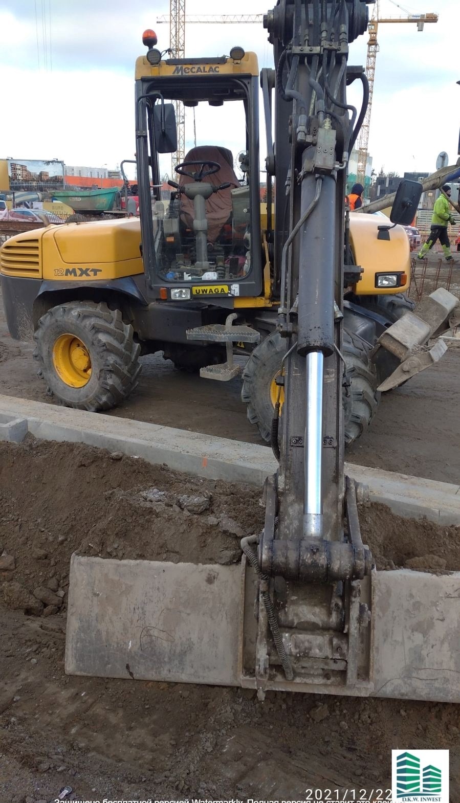 Żółta koparko-ładowarka Mecalac 12MXT z łyżką zagłębiającą się w ziemię na placu budowy, widoczne elementy betonowe i żuraw budowlany w tle.