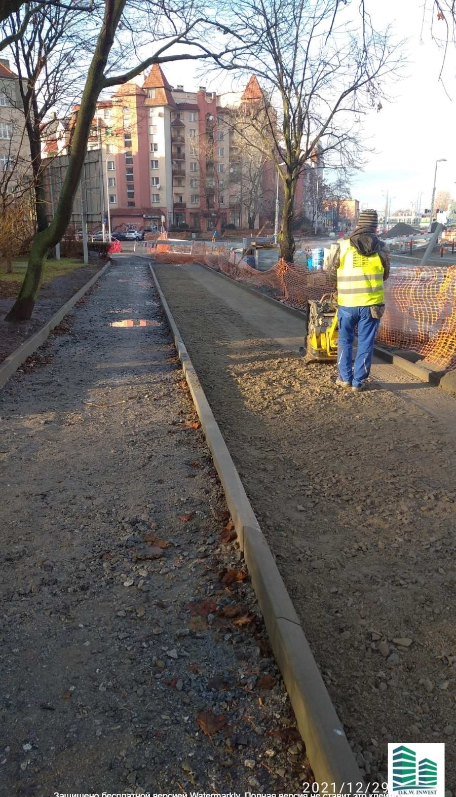Pracownik w kamizelce odblaskowej zagęszcza podłoże pod chodnik za pomocą zagęszczarki wibracyjnej, w tle budynki mieszkalne i drzewa.