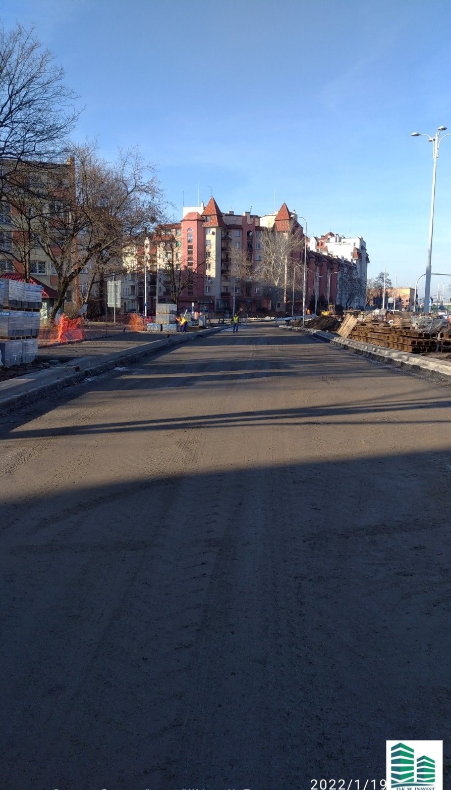 Nowa nawierzchnia asfaltowa na miejskiej ulicy w trakcie budowy, z widocznymi robotnikami w oddali i budynkami mieszkalnymi w tle.