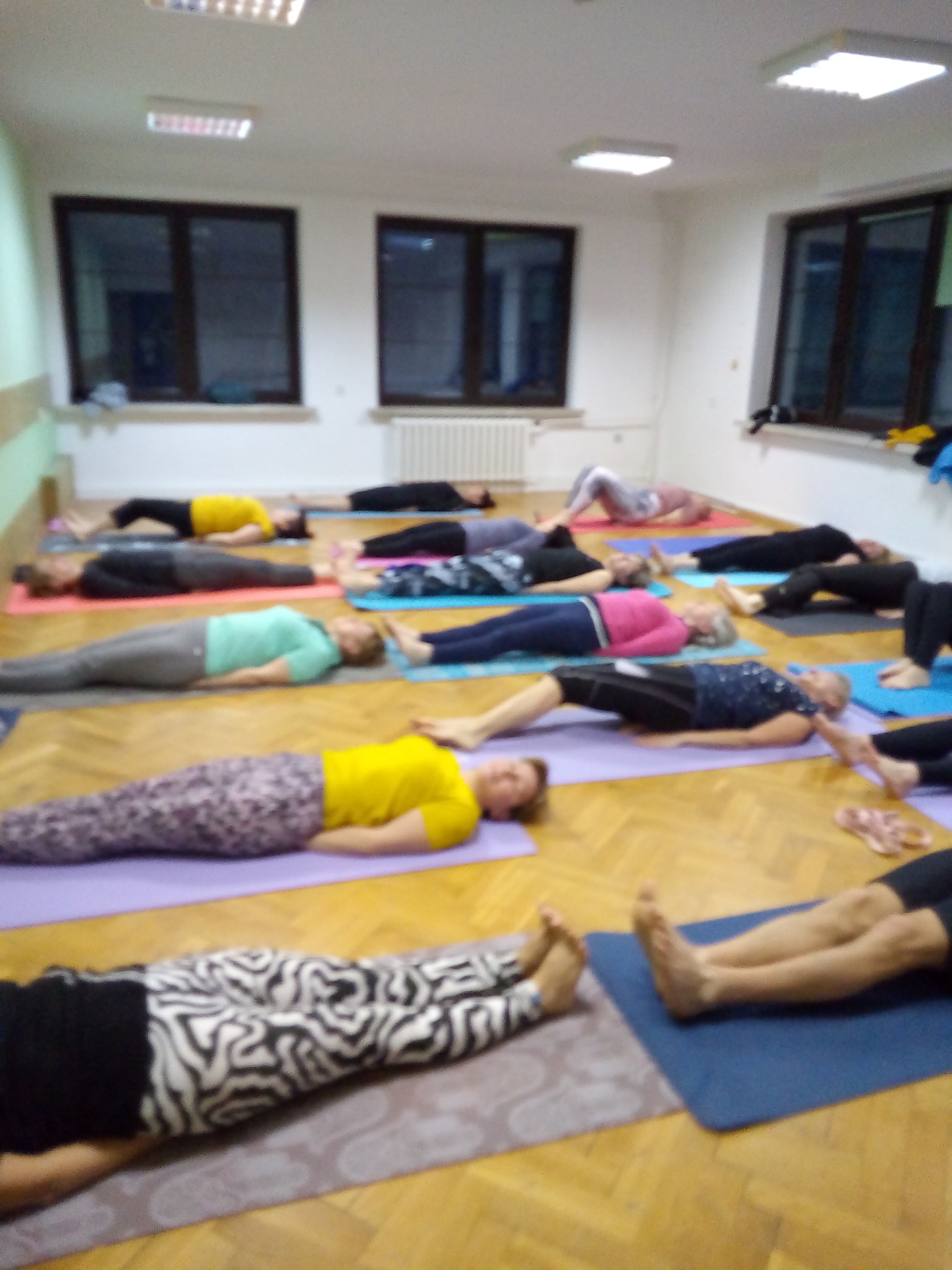 Grupa osób relaksuje się na matach podczas warsztatów w Częstochowie, leżąc na plecach w sali z parkietem i oknami.