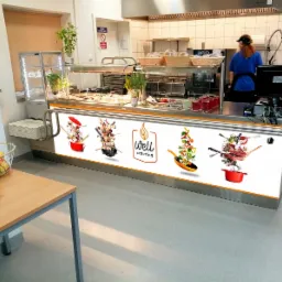 Bufet z ekspozycją dań i dekoracyjnym panelem z logo 'well catering' oraz unoszącymi się warzywami i przyborami kuchennymi, w tle pracująca osoba w kuchni.