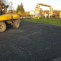 Żółta koparka Mecalac 12MXT na utwardzonym terenie z widocznymi śladami opon, w tle budynki i reklamy firm.