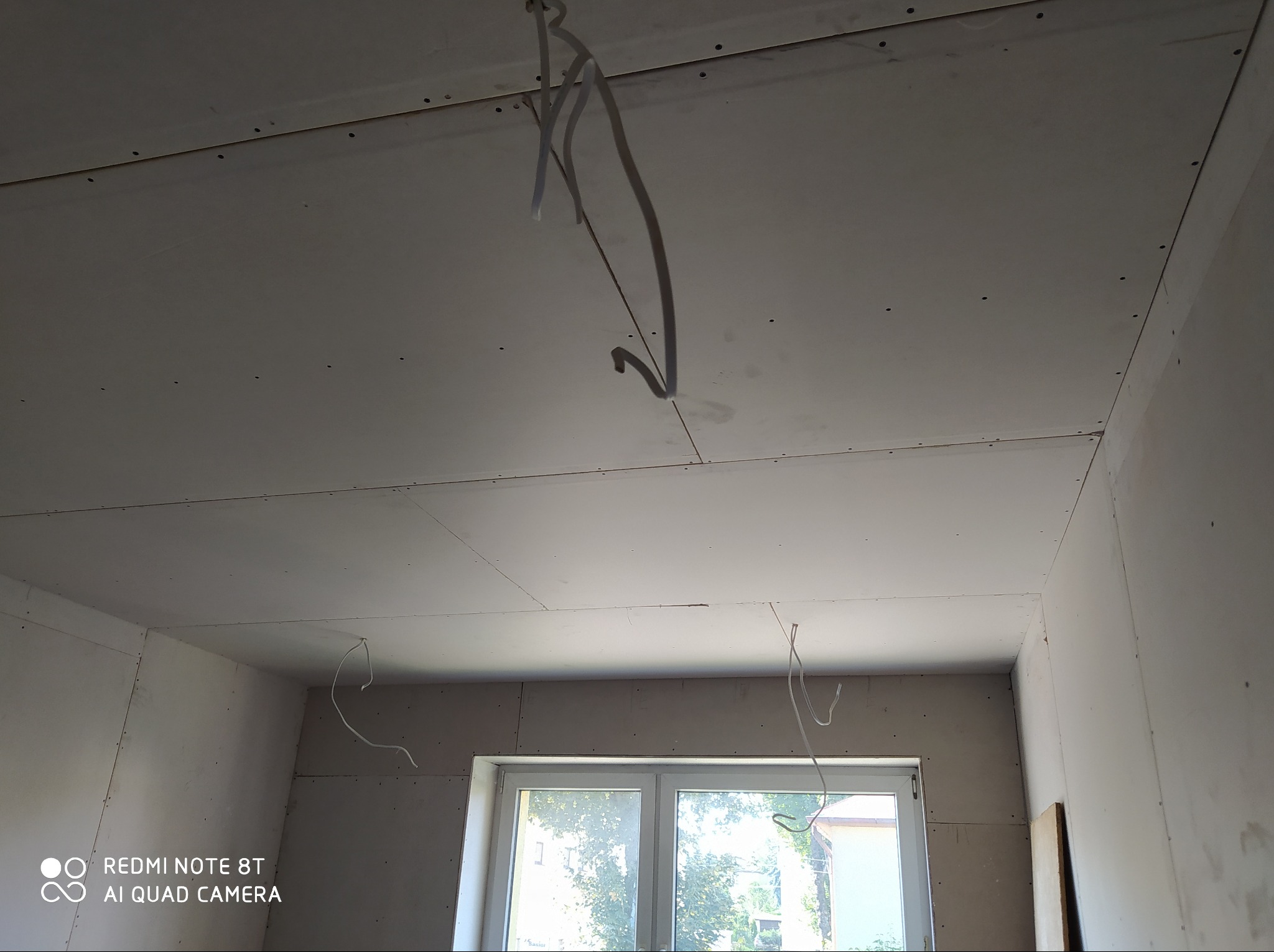 Wnętrze pokoju w trakcie remontu z zamontowanymi płytami na suficie i ścianach, widoczne przewody elektryczne zwisające z sufitu, okno z widokiem na zieleń na zewnątrz.