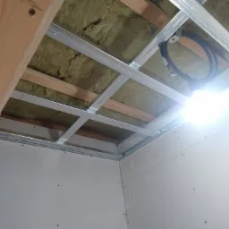 Sufit w trakcie montażu: widoczne profile metalowe tworzące konstrukcję, izolacja z wełny mineralnej oraz częściowo zamontowane płyty gipsowo-kartonowe na ścianach. W tle widoczne drewniane...