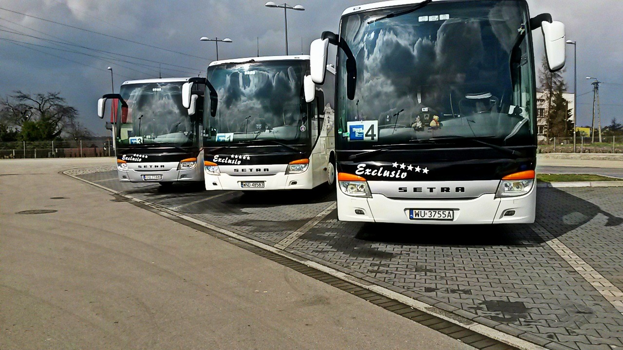 Trzy białe autokary Setra Exclusivo zaparkowane na parkingu z kostki brukowej, widok z przodu, pochmurne niebo odbija się w szybach.