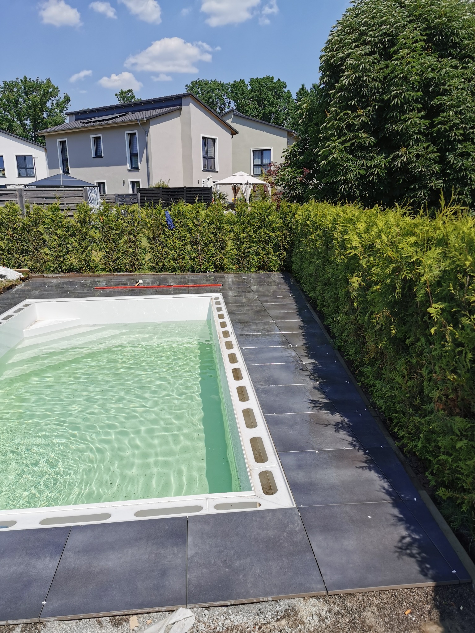 Napełniony wodą basen z białym obramowaniem i ułożoną ciemnoszarą kostką brukową wokół, na tle zielonego żywopłotu i domów.