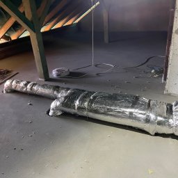 Somergy Sp. z o.o. - Izolowany przewód wentylacyjny na poddaszu, widoczne elementy konstrukcji dachu i instalacje elektryczne. Szare podłoże, surowy stan wykończenia.