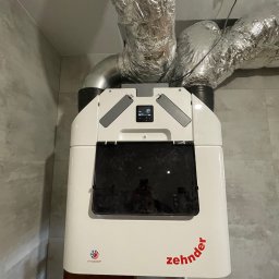 Somergy Sp. z o.o. - Centrala rekuperacyjna Zehnder z podłączonymi przewodami wentylacyjnymi w pomieszczeniu z szarą płytką. Widoczny panel sterowania i logo producenta.