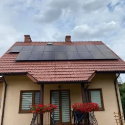 Dom jednorodzinny z panelami słonecznymi na czerwonym dachu, widok z przodu, balkon z czerwonymi kwiatami, pochmurne niebo w tle.