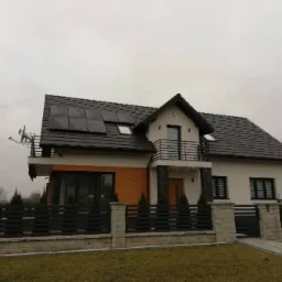 Nowoczesny dom murowany z panelami fotowoltaicznymi na dachu, czarną balustradą balkonu, drewnianą elewacją i ogrodzeniem z szarego kamienia.