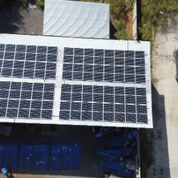 Widok z góry na panele słoneczne zamontowane na dachu budynku przemysłowego, obok zaparkowany samochód i składowane niebieskie plandeki.