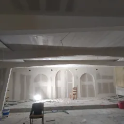 Wnętrze w trakcie prac wykończeniowych: ściany z elementami łukowymi przygotowane pod gładzie, widoczne płyty kartonowo-gipsowe, dwa krzesła, wiadra z farbą i przewody elektryczne. Na podłodze...