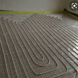 Montaż ogrzewania podłogowego: białe rurki ułożone w pętle na folii z czerwoną siatką, widoczne kable i taśma zabezpieczająca przy ścianach.