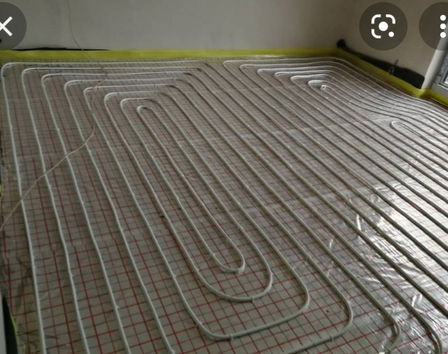 Montaż ogrzewania podłogowego: białe rurki ułożone w pętle na folii z czerwoną siatką, widoczne kable i taśma zabezpieczająca przy ścianach.