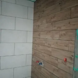 Ściana łazienki wyłożona białymi prostokątnymi płytkami oraz drewnopodobnymi panelami, widoczne otwory na instalacje wodne i fragment ściany z zielonym gruntem, etap prac wykończeniowych.