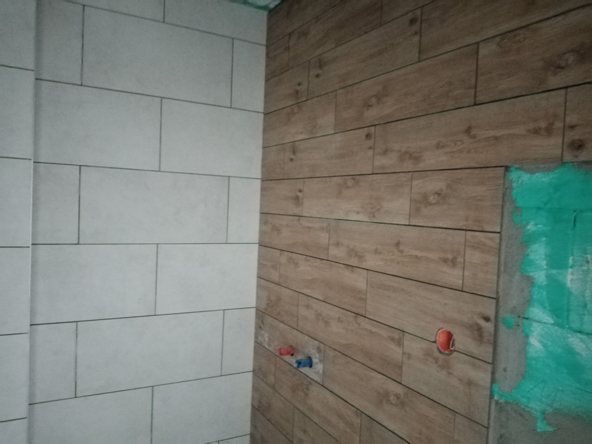 Ściana łazienki wyłożona białymi prostokątnymi płytkami oraz drewnopodobnymi panelami, widoczne otwory na instalacje wodne i fragment ściany z zielonym gruntem, etap prac wykończeniowych.