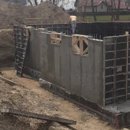 Wykop pod fundamenty z częściowo zabetonowanymi ścianami szalunkowymi, widoczne drewniane konstrukcje okienne i drabina oparta o ścianę, w tle dom z czerwonym dachem i drzewa bez liści.