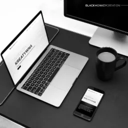 Czarno-białe zdjęcie biurka z laptopem, smartfonem, kubkiem kawy i monitorem. Na ekranie laptopa i smartfona wyświetlona strona internetowa agencji kreatywnej BlackMonkeyCreation.