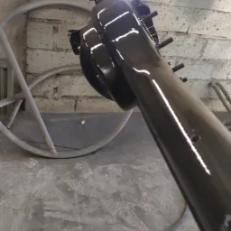 Błyszczący, czarny element metalowy po malowaniu w Dobczycach, widoczne refleksy światła, w tle szary mur z cegieł i fragmenty ramy, na podłodze brudna szmatka i przewód.