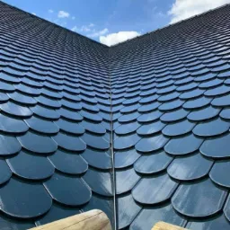 Ukończony dach pokryty ciemnymi, błyszczącymi dachówkami w kształcie łuski, z widocznym fragmentem drewnianego rusztowania i błękitnym niebem w tle.