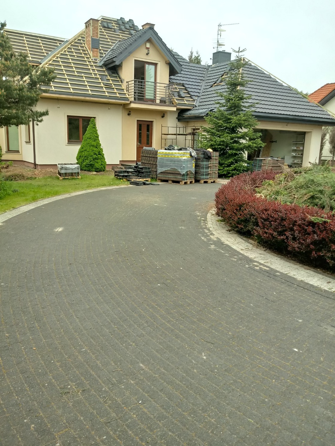 Dom jednorodzinny w trakcie prac dekarskich; widoczne łatowanie dachu, częściowo pokryty dachówką, materiały budowlane na paletach przed budynkiem.