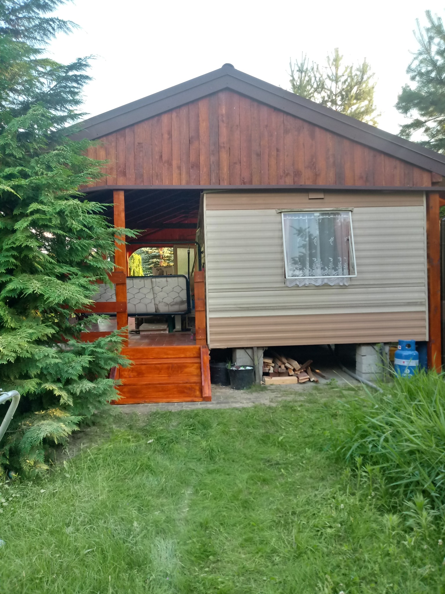 Domek letniskowy z drewnianą werandą i schodami, widoczne butle gazowe i drewno pod spodem, otoczony zielenią.