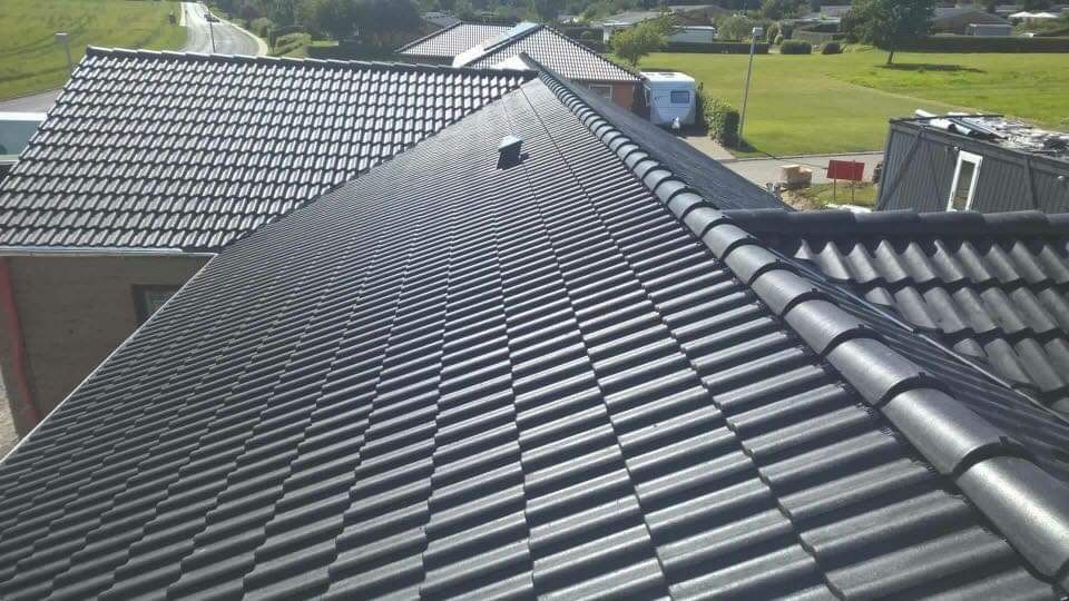 Nowy dach z ciemnej dachówki ceramicznej, widok z góry na gotowy dach z kominkiem wentylacyjnym, w tle zabudowa jednorodzinna i zieleń.