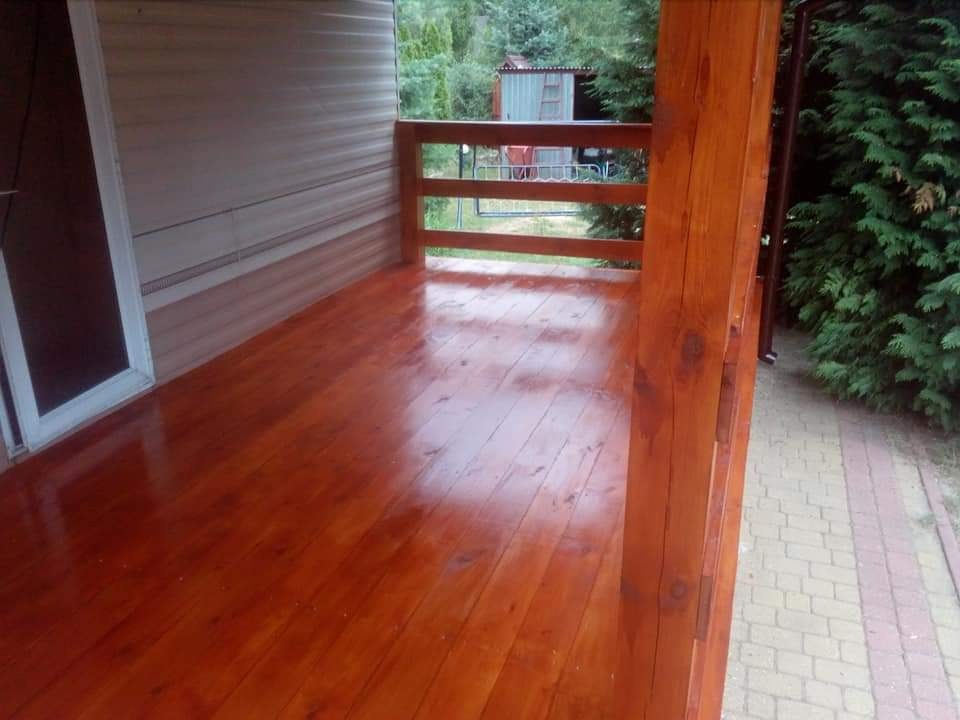 Błyszcząca, świeżo polakierowana drewniana weranda z balustradą, widok z boku, z elementami budynku w tle i zielenią.