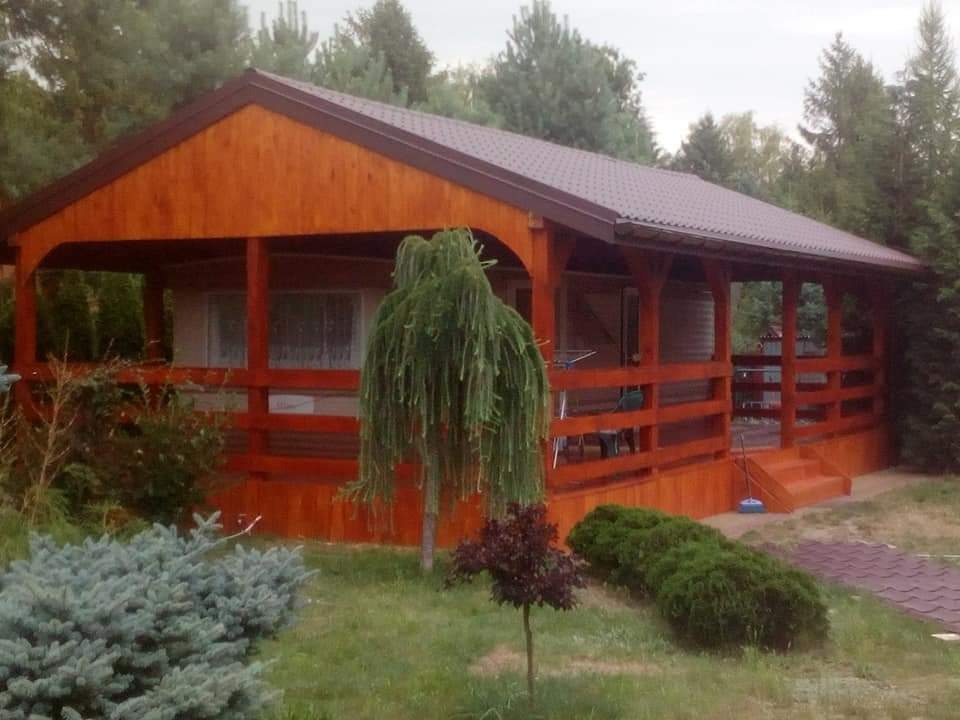 Drewniana altana z brązowym dachem w ogrodzie, widoczne schody, balustrada i drzewka ozdobne.