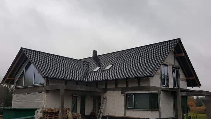 Dom w trakcie budowy z nowym, ciemnym dachem z dachówki, widoczne okna dachowe i komin, surowy stan elewacji.