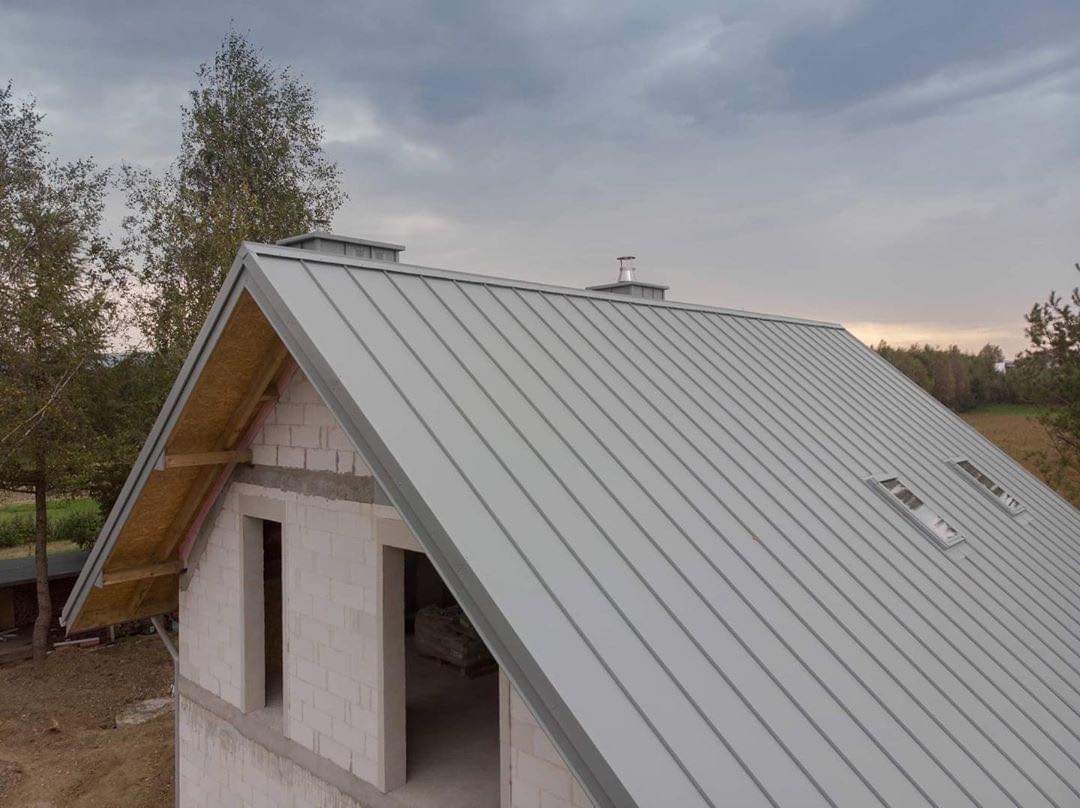 Szary dach z blachy na surowym budynku w trakcie budowy, widoczne okna dachowe i komin, w tle drzewa i pochmurne niebo.