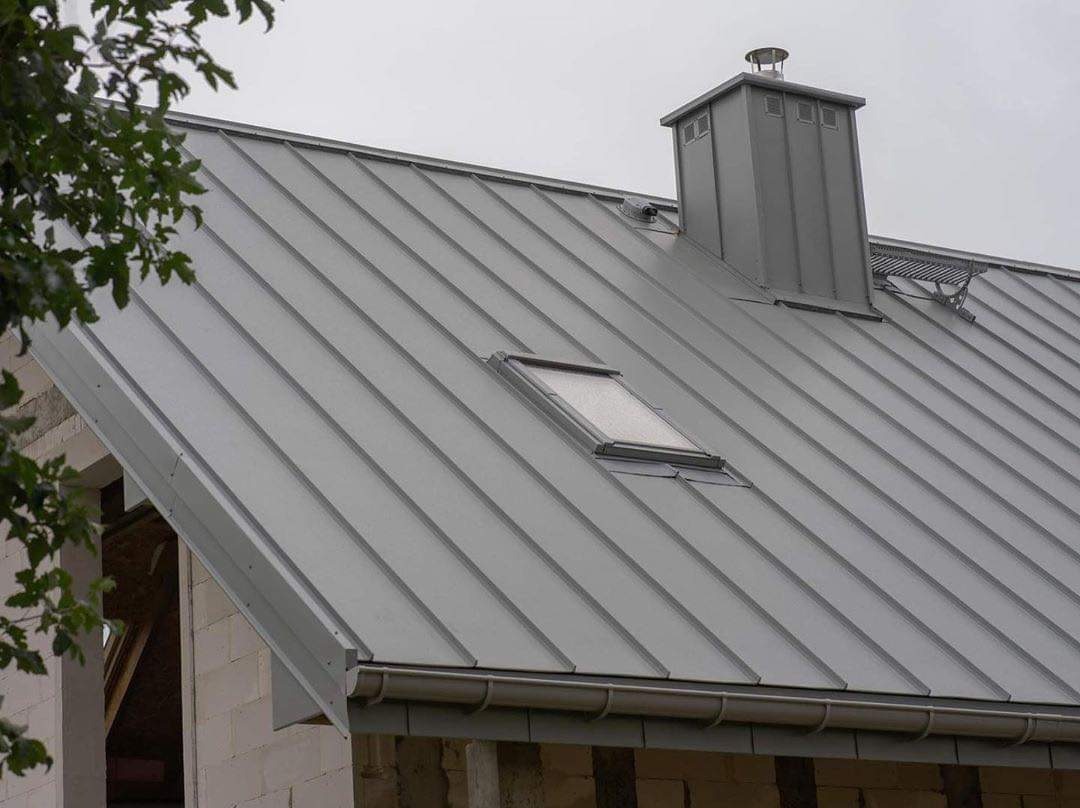 Szary dach z blachy na surowym budynku z oknem dachowym i kominem, widoczne rynny.