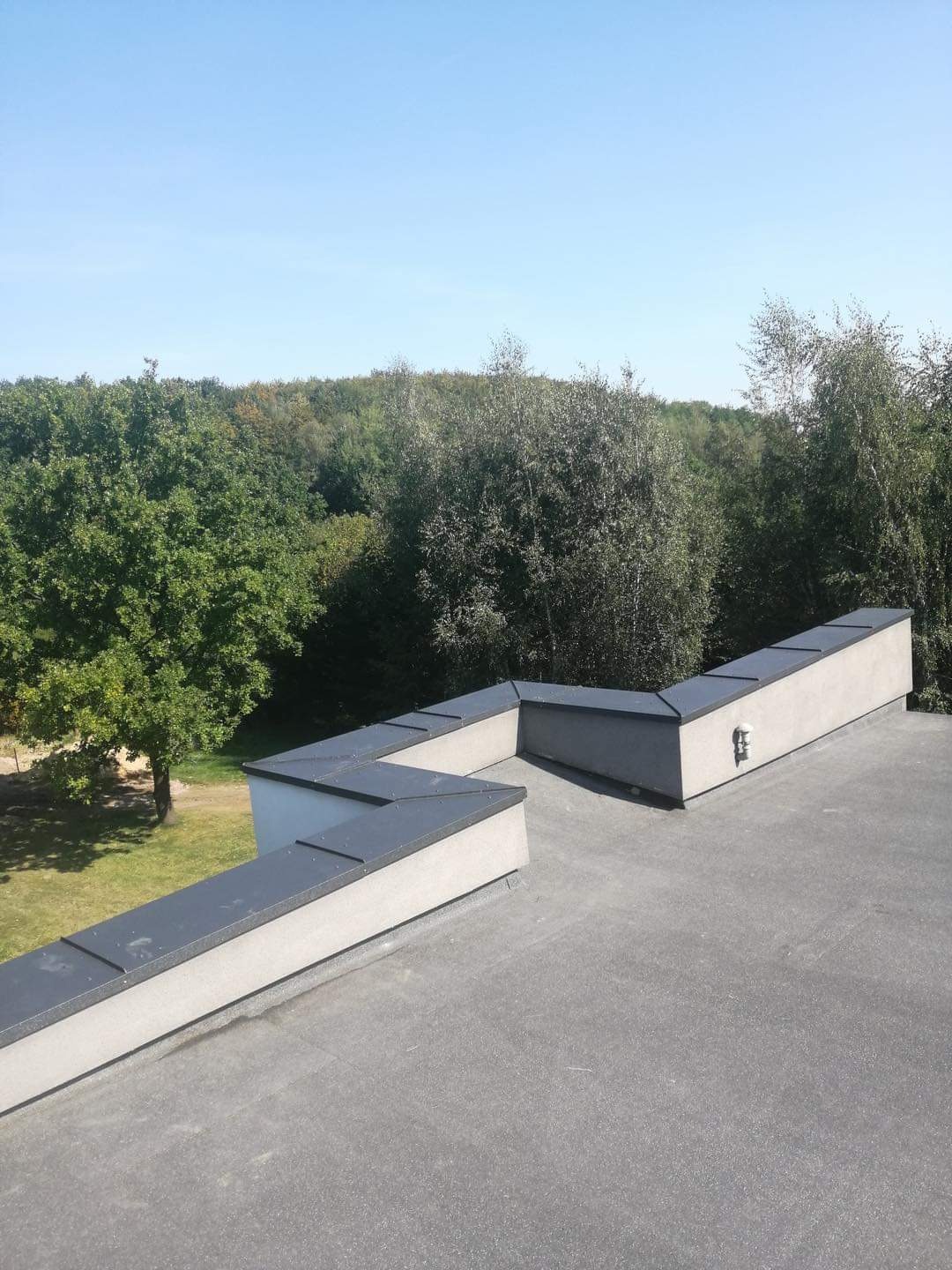 Płaski dach pokryty papą termozgrzewalną z attyką i obróbkami blacharskimi, widok z góry na tle zieleni drzew.