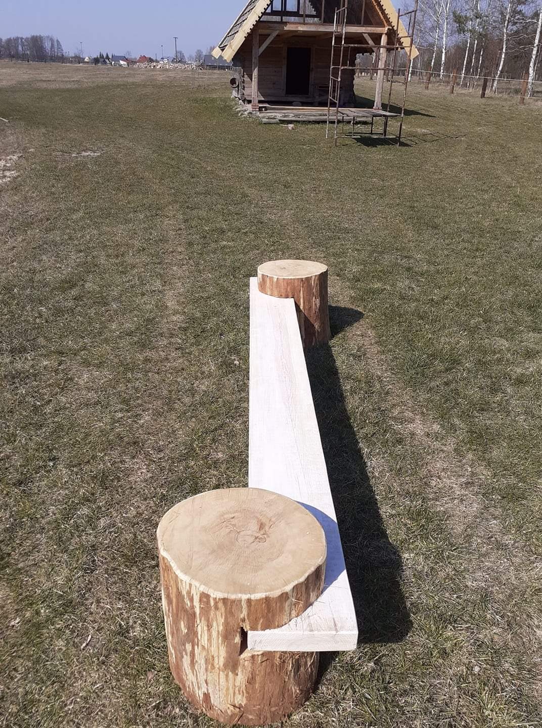 Surowe drewniane siedzisko z bali i deski na trawie, w tle częściowo wykończona konstrukcja drewniana przypominająca budynek z dachem w trakcie budowy.