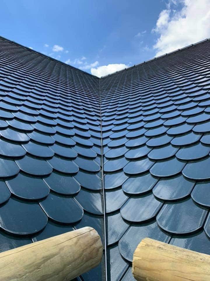 Ukończony dach pokryty ciemnymi, błyszczącymi dachówkami w kształcie łuski, z widocznym fragmentem drewnianego rusztowania i błękitnym niebem w tle.