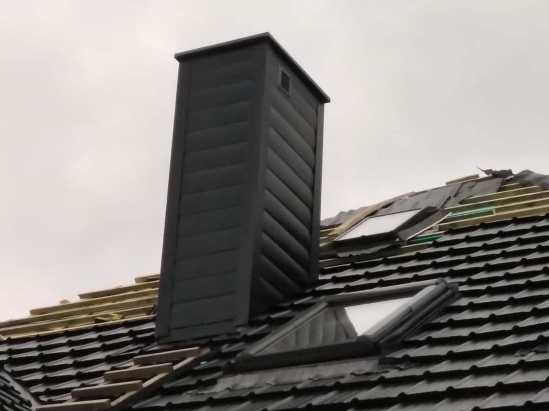 Ciemny komin wentylacyjny z żaluzjami na tle fragmentu dachu z czarną dachówką, widoczne okna dachowe i elementy konstrukcji dachu w trakcie prac.