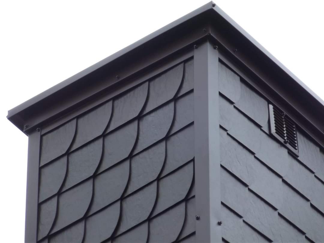 Szczegółowe ujęcie fragmentu dachu pokrytego ciemnoszarymi, łukowatymi płytkami łupek, z widoczną kratką wentylacyjną i metalowym obramowaniem na krawędzi.