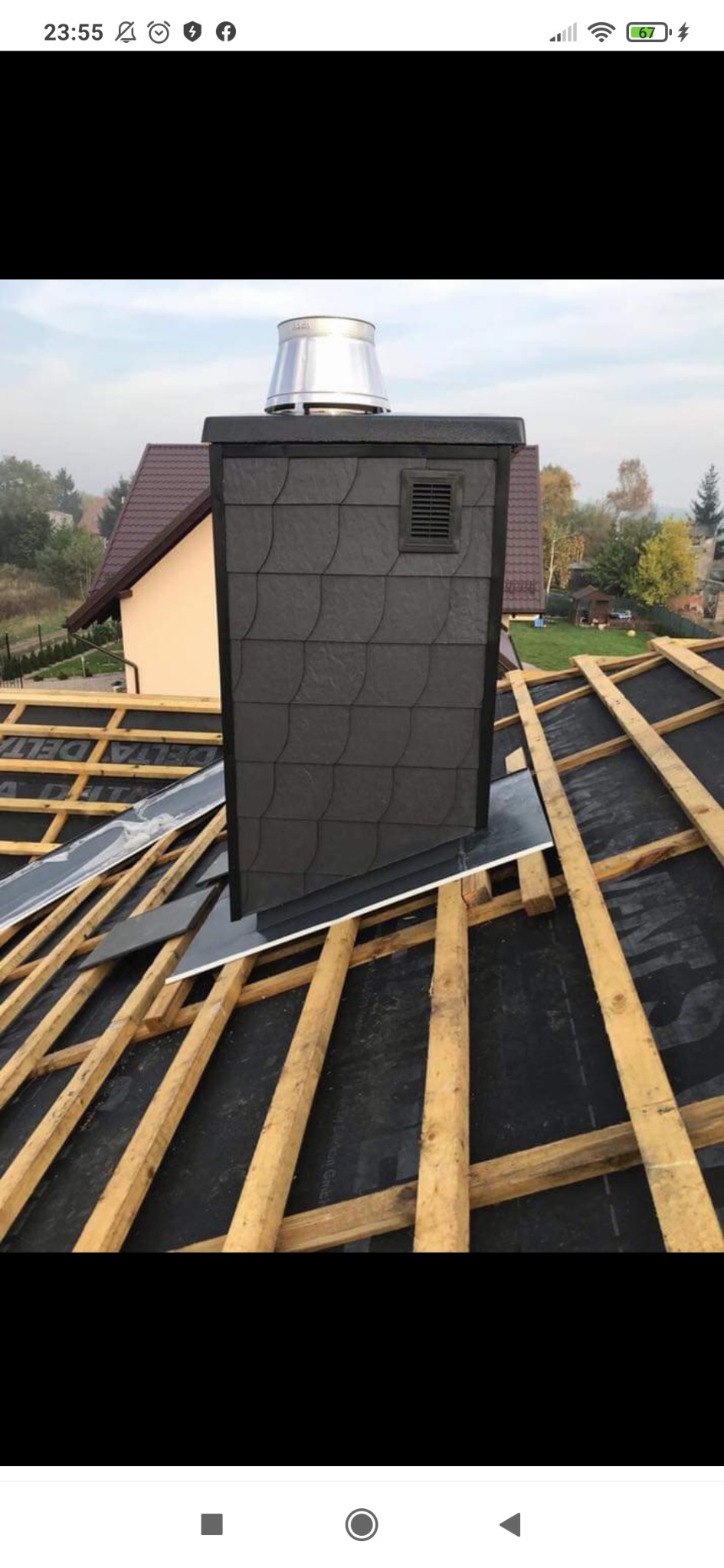 Kominek wentylacyjny z szarej łupki na dachu w trakcie budowy, widoczne drewniane krokwie i membrana dachowa, w tle zabudowa jednorodzinna.