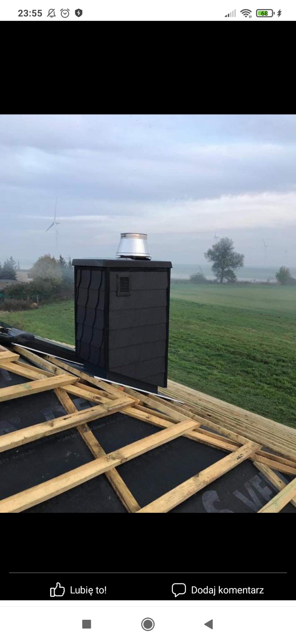 Ciemnoszary komin z metalowym daszkiem na tle zielonego pola i wiatraków, widoczny szkielet konstrukcji dachu pokryty czarną membraną.