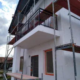 Dom w trakcie ocieplania, widoczne rusztowania, styropianowe płyty elewacyjne, zabezpieczone okna i drzwi, balkon z czarną balustradą.