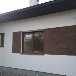 Fragment elewacji domu z białym tynkiem i brązowymi panelami imitującymi drewno, brązowe okna z roletami, rynna w kolorze elewacji.