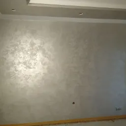 Ściana wewnętrzna pokryta dekoracyjnym tynkiem w odcieniach szarości i srebra, z widocznymi gniazdkami elektrycznymi i listwą przypodłogową z jasnego drewna.