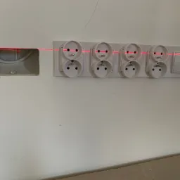 Biała ściana z zamontowanymi podwójnymi gniazdkami elektrycznymi i włącznikiem, wyrównanymi poziomą wiązką lasera; po lewej widoczne przygotowane miejsce na kolejne gniazdko z wystającymi kablami.